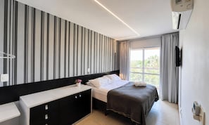Una habitación de hotel moderna con una pared a rayas, una cama con cabecera oscura, una mesita de noche con un jarrón de flores y un ventanal con cortinas.