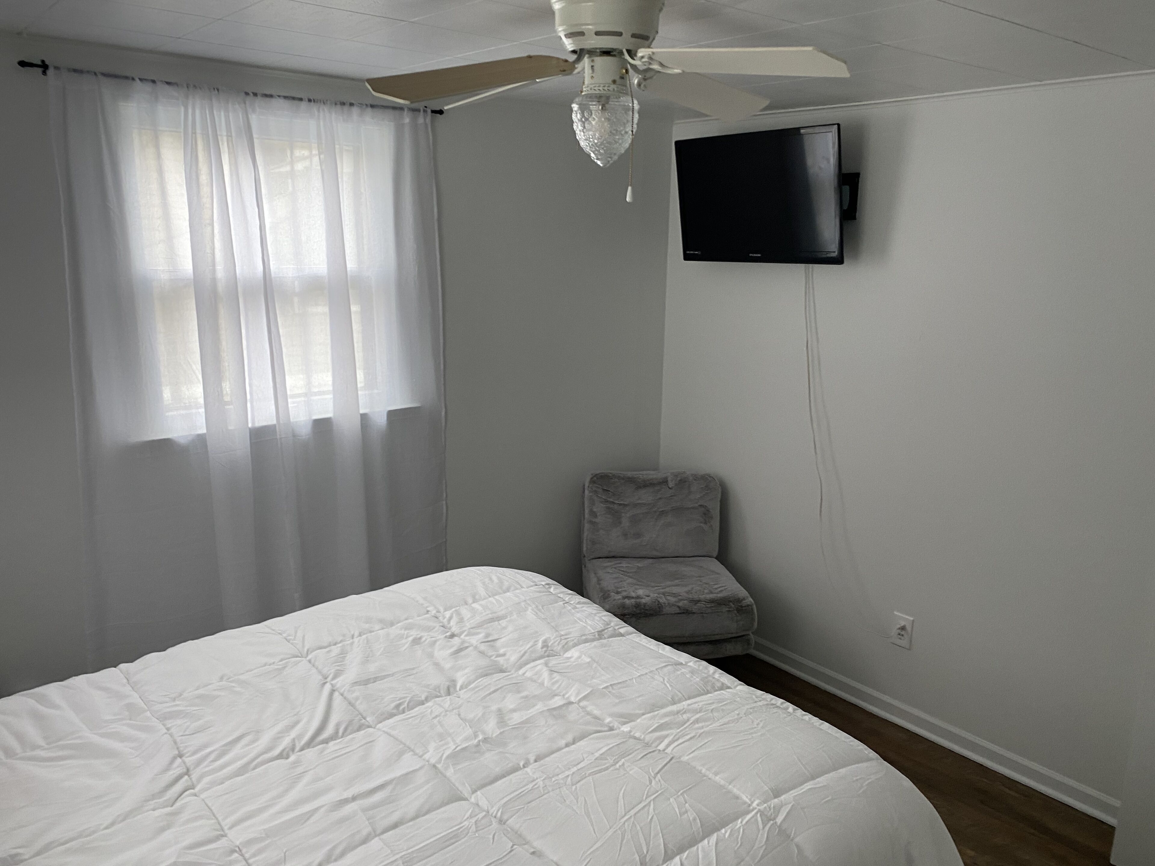 Un dormitorio con cama, ventilador de techo, televisor montado en la pared y una silla.