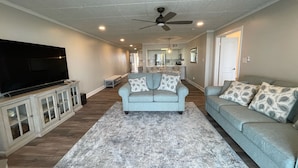 A spacious living room with a large flat-screen TV, a ceiling fan, and a light-colored carpet.