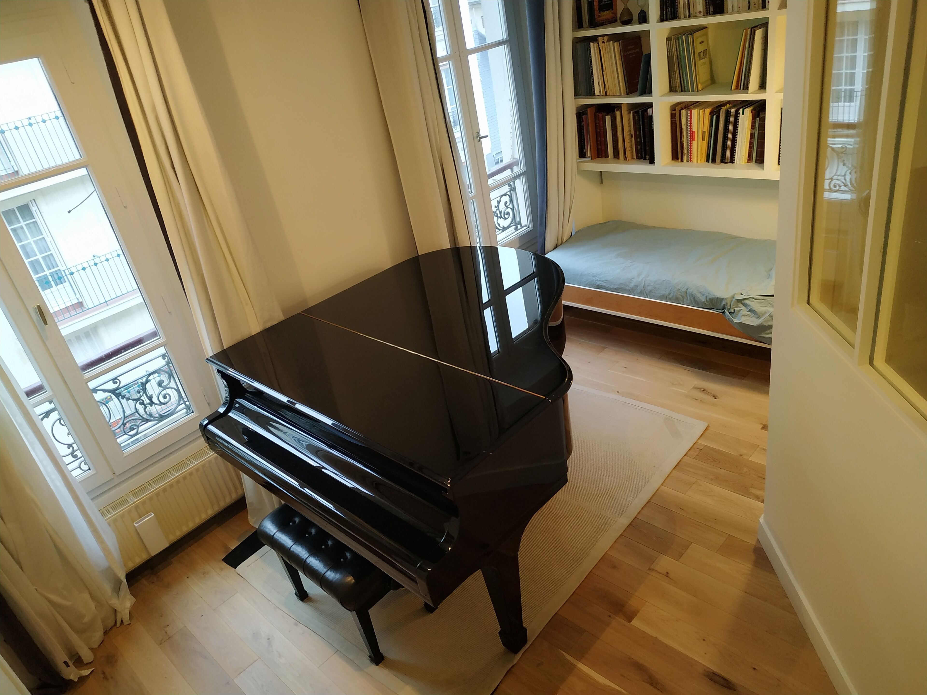 Una habitación con un piano, una cama, una estantería y una ventana con vistas a un balcón.