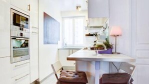 A modern kitchen with white cabinets, a built-in oven, a dining table with two stools, and a pink lamp.