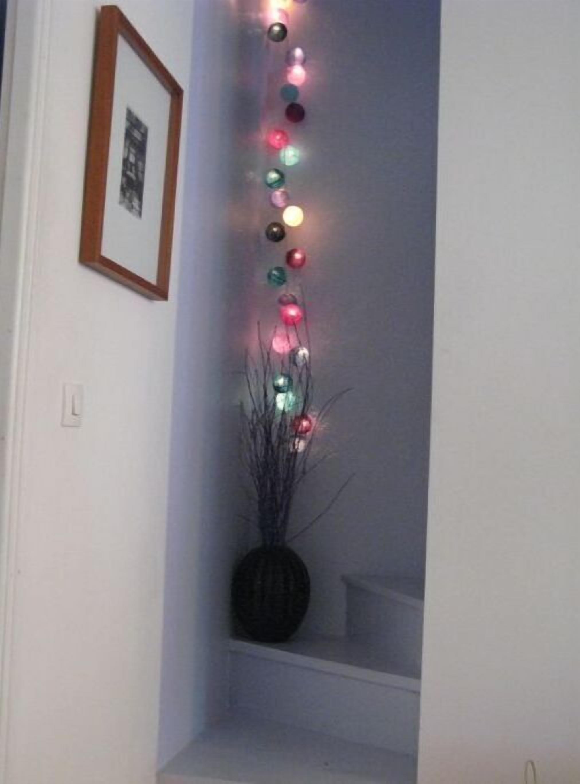Un coin de pièce avec un mur blanc, un tableau encadré et une petite étagère sur laquelle se trouvent un vase et des guirlandes lumineuses décoratives.
