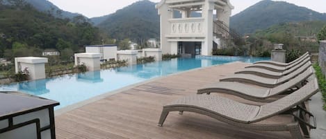 A poolside area with lounge chairs, a white building with a peaked roof, and a mountainous backdrop.