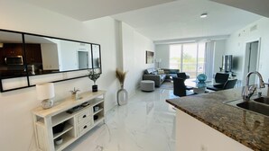 A modern kitchen with a marble countertop, a dining area with a glass table, and a living room with a sofa and a TV. - Serenity Ocean Luxury Condo (Boynton Beach)
