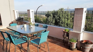 Una terraza con una mesa redonda y sillas, plantas en macetas y vistas a árboles y montañas a lo lejos.