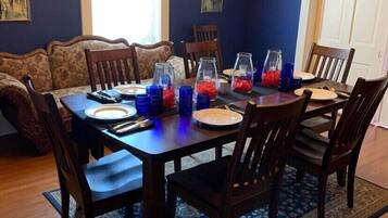 Comedor con mesa de madera dispuesta para cuatro personas, rodeada de sillas, con una araña colgante y un tapete estampado en el suelo.