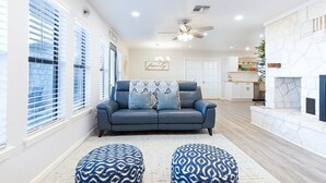 A spacious living room with a blue sofa, patterned ottomans, a ceiling fan, and a stone fireplace.