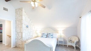 A bedroom with a stone wall, a bed with white bedding, two bedside tables with lamps, a chair, and a ceiling fan.