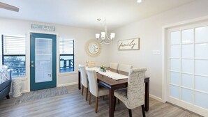 A dining area with a wooden table, white chairs, a chandelier, and a 'family' sign on the wall.