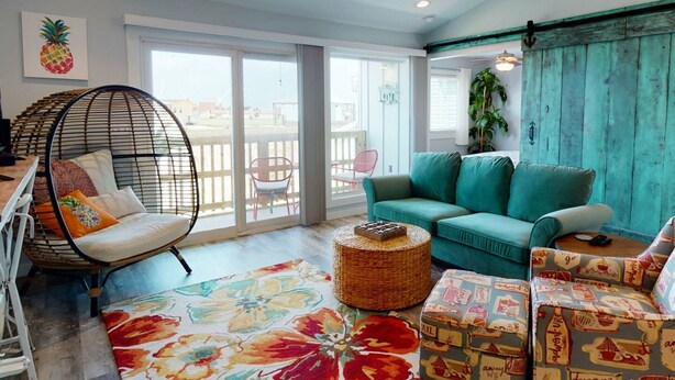 Een ruime woonkamer met een groot raam, een hangstoel, een blauwgroene bank en een kleurrijk vloerkleed.