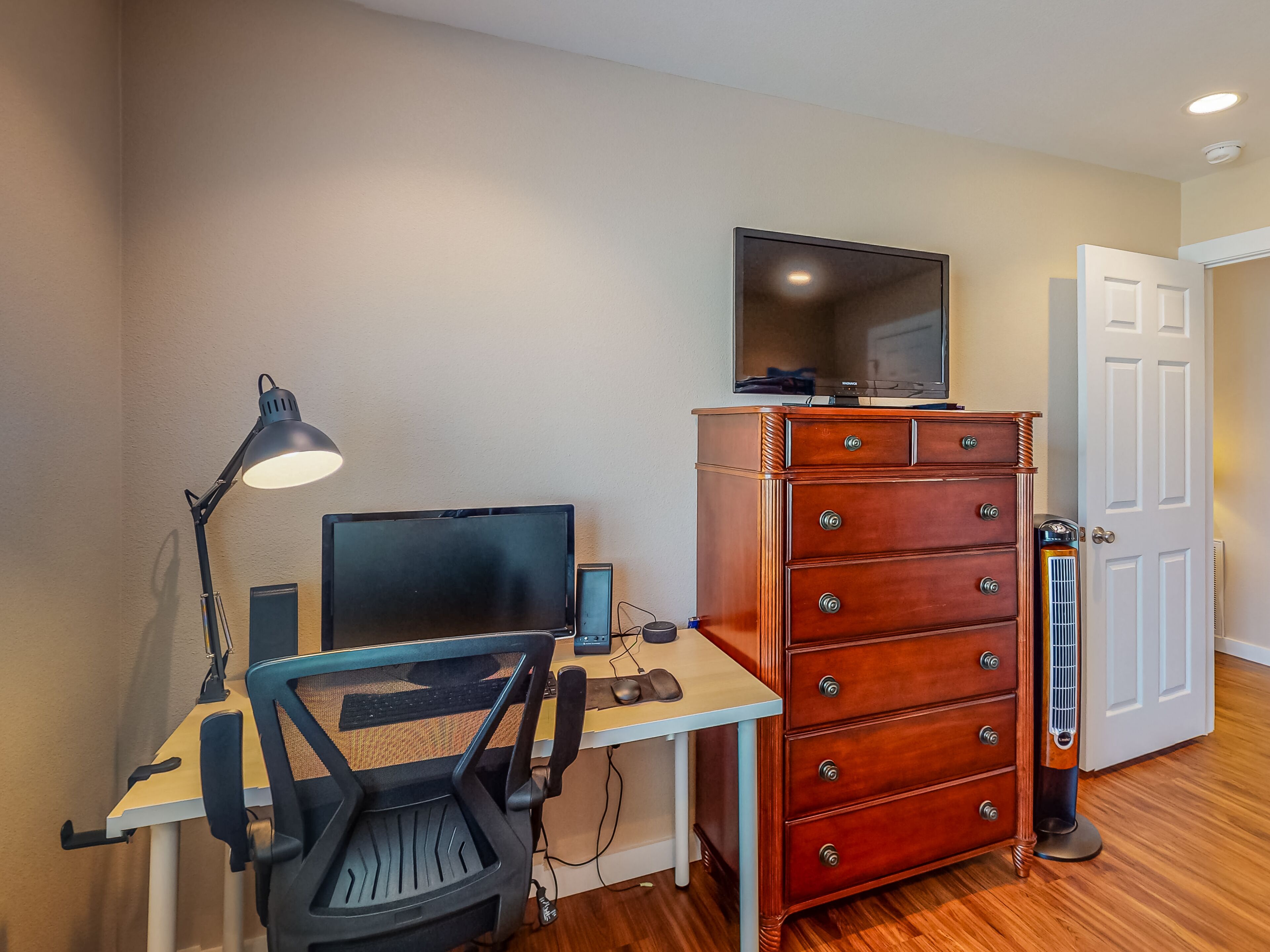 Ein kleiner Arbeitsraum mit Schreibtisch, Computer, Stuhl, Lampe, Kommode und Fernseher an der Wand.