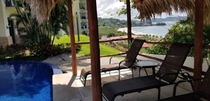 A balcony with a pool, chairs, and a view of a beach and mountains.