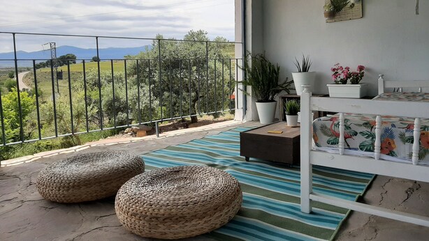 En balkon med udsigt over landskabet, en hvid seng, fletpuder og plantede planter.