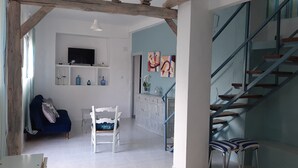 En rummelig opholdsstue med sofa, hvid stol og spisebord. En trappe med blå gelænder fører til en etage ovenpå.