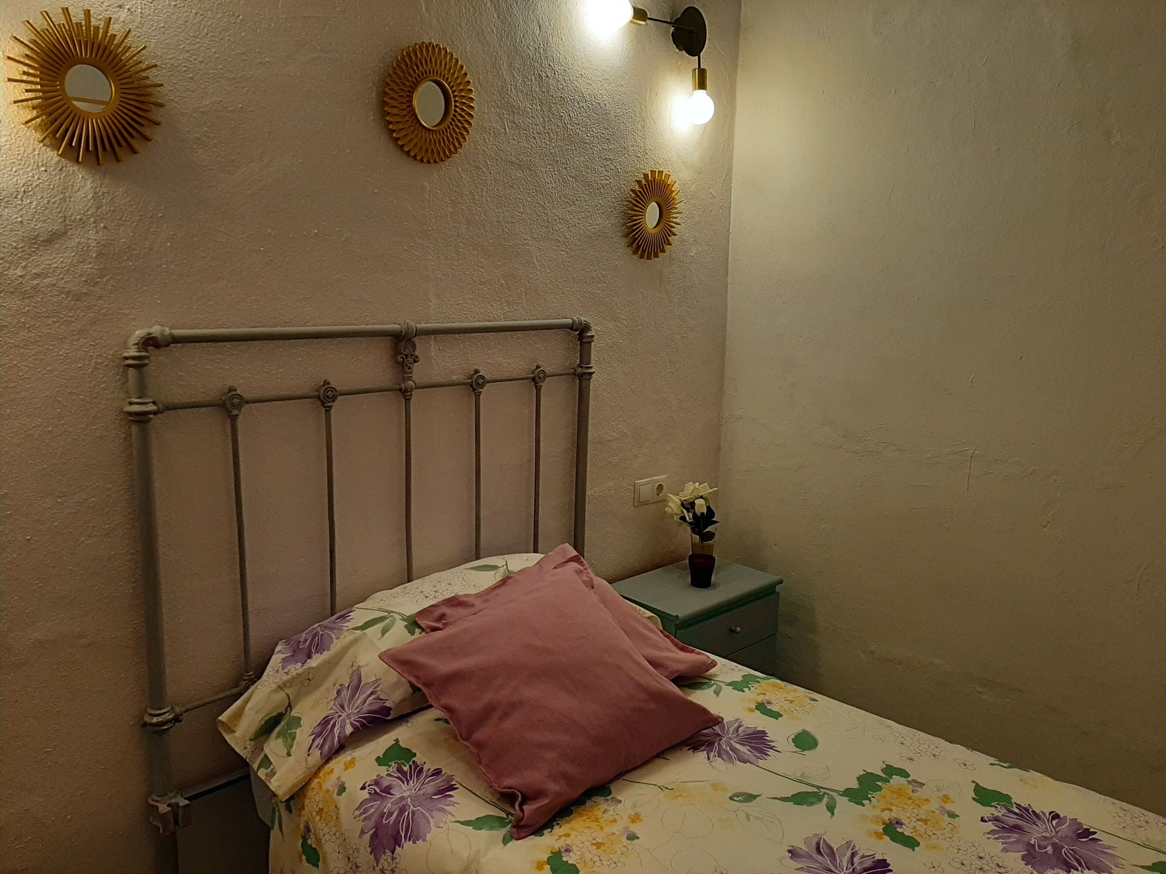 Een bed met een metalen hoofdbord, een bloemenbedekking en één roze kussen. Er zijn wandlampen, een kleine tafel met een bloevas en twee zonneschijf spiegelwanden.