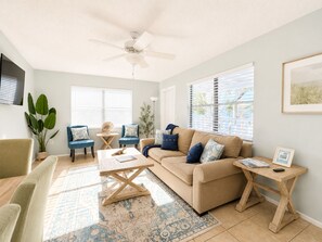 Un salon avec un canapé beige, des fauteuils bleus, une table basse en bois, un ventilateur de plafond et une grande fenêtre avec des stores.