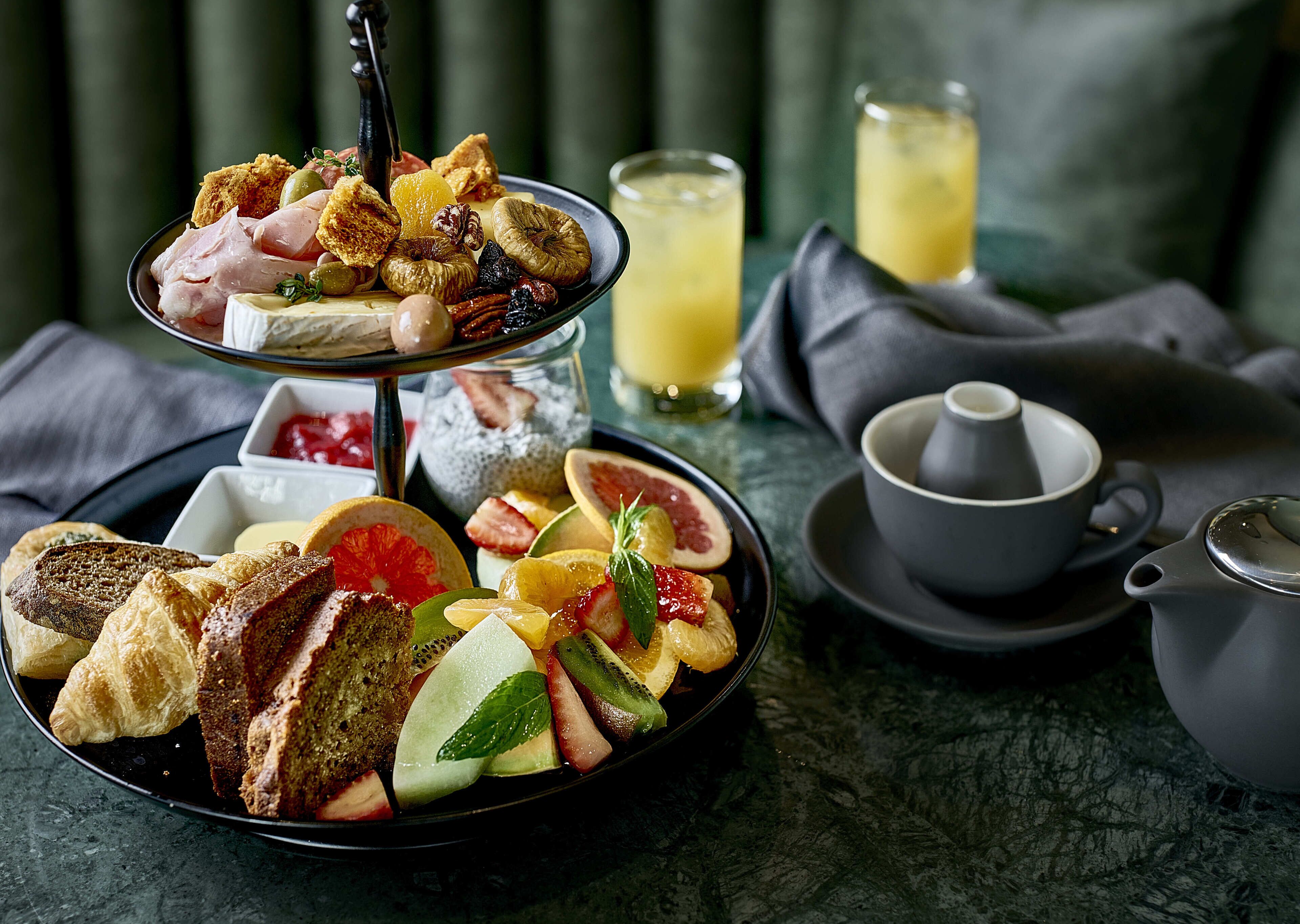 A breakfast setting with a three-tiered fruit and pastry plate, a glass of orange juice, and a teapot on a marble table.