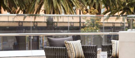 A balcony with cushioned seating and a small table, overlooking palm trees and a building.