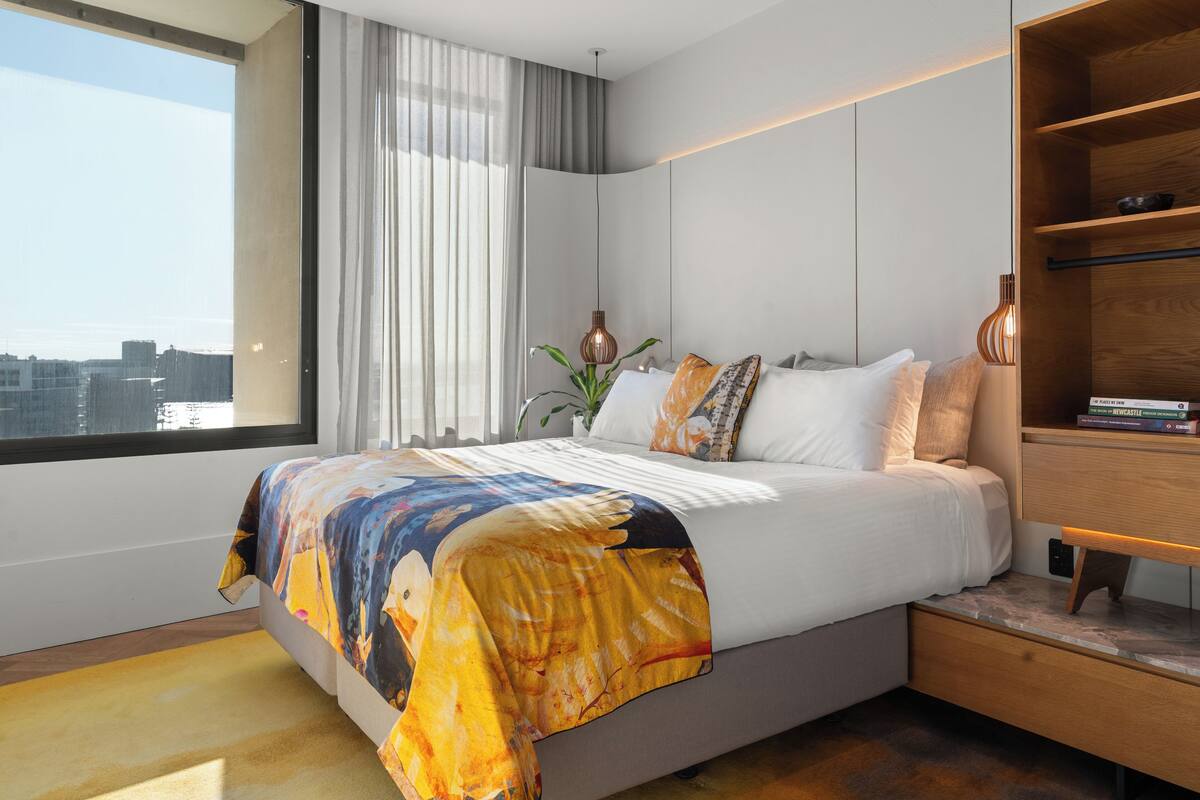 A modern bedroom with a large bed, a wooden nightstand, and a view of the city through a large window.