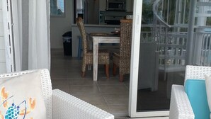 Un soggiorno moderno con mobili in vimini bianco, una porta scorrevole in vetro che dà su un balcone, e vista sulla cucina e zona pranzo.