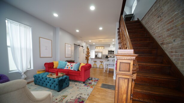 A spacious living room with a red sofa, blue ottoman, and a colorful rug. A wooden staircase leads to an upper level.