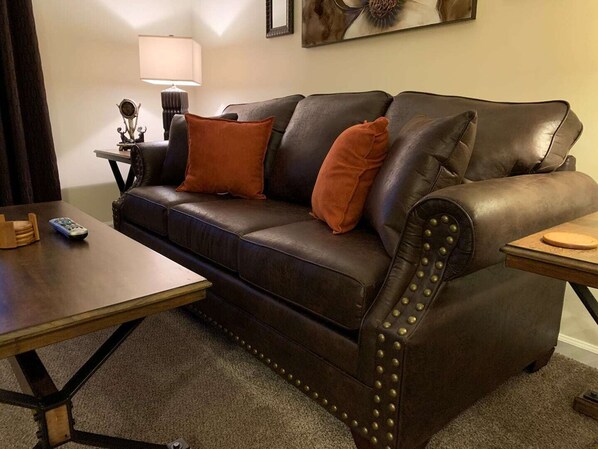Un canapé en cuir marron foncé avec des coussins orange, une petite table avec une télécommande et une lampe sur une table d’appoint.
