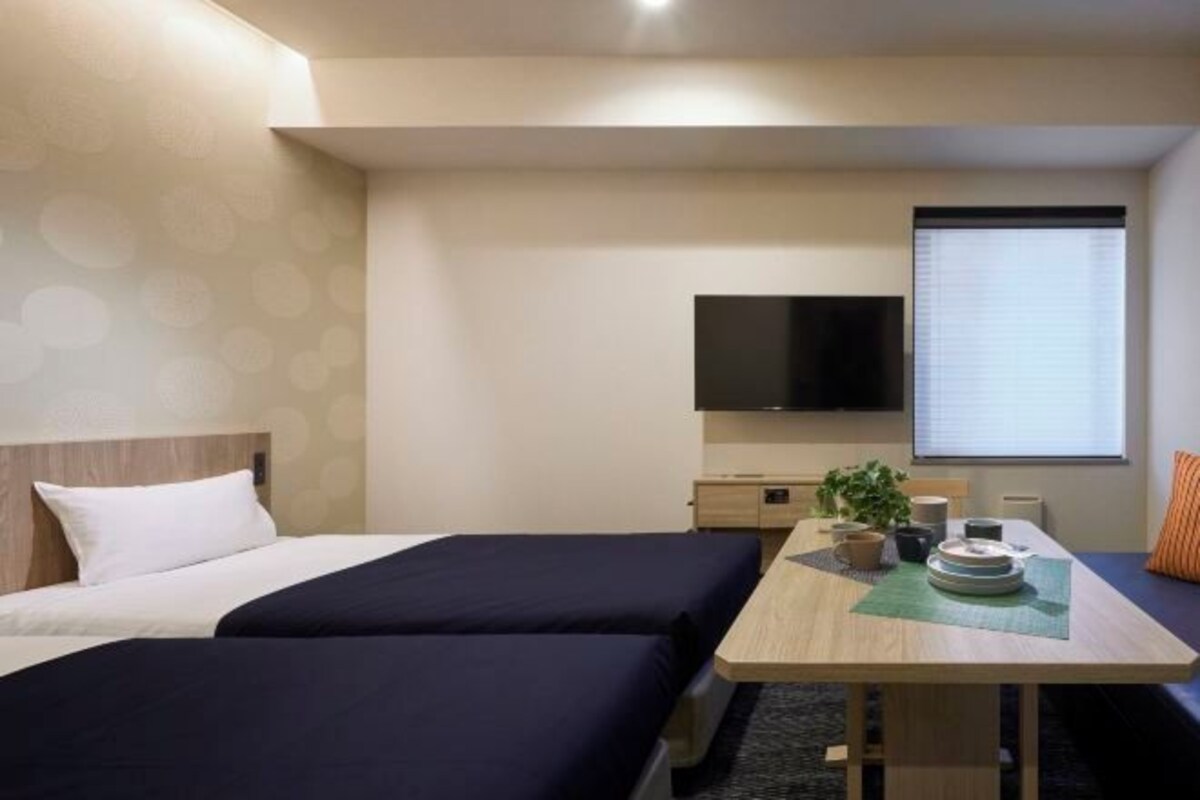 A hotel room with two beds, a flat-screen TV, a small table with a plant, and a window with blinds.