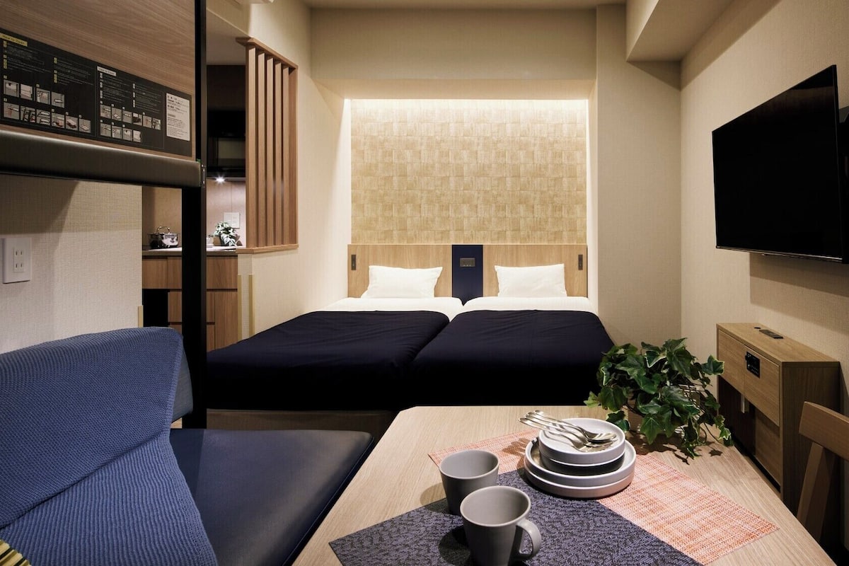 Habitación de hotel compacta con literas, un sofá, una mesa de comedor con una planta y un televisor de pantalla plana.