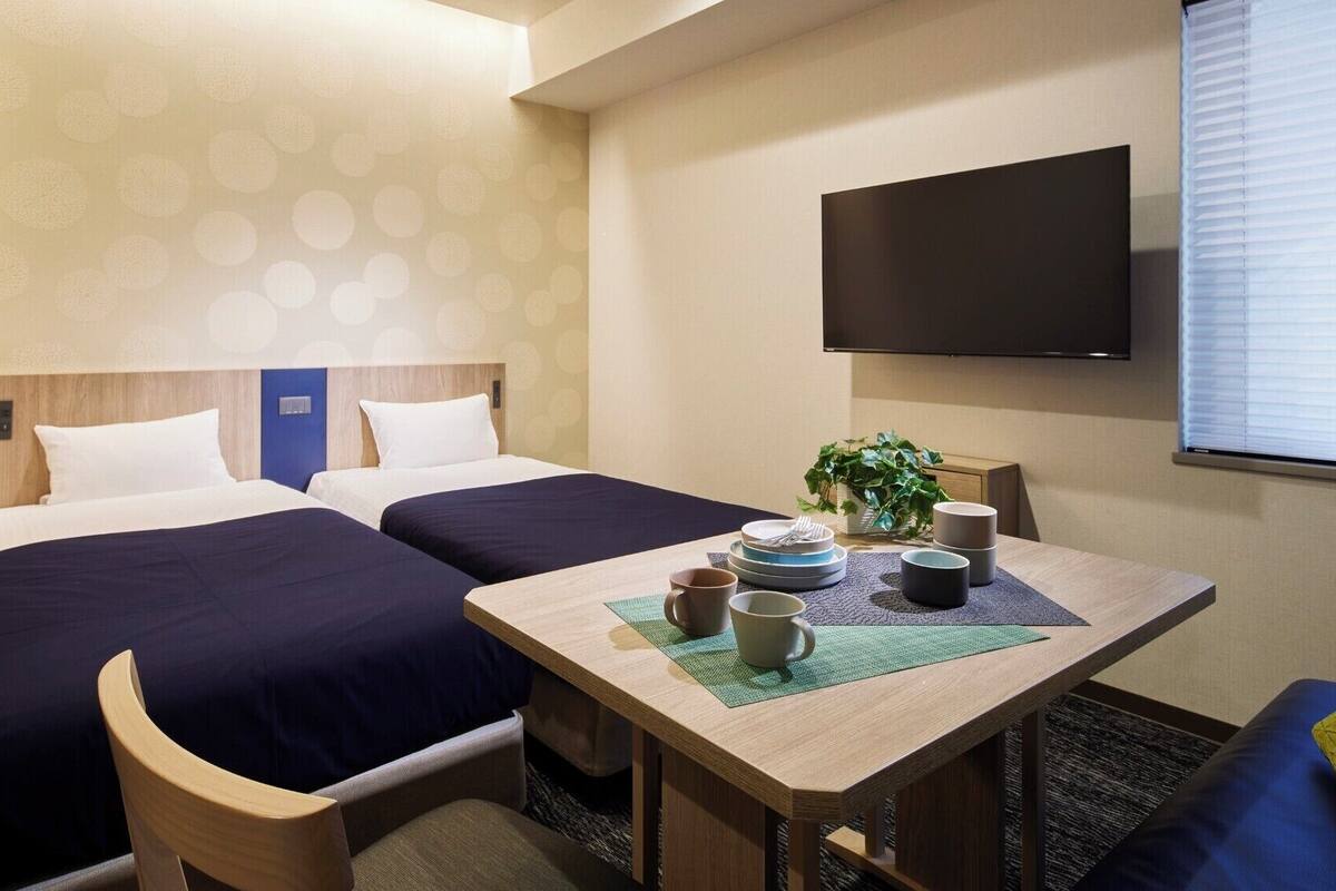 Una habitación de hotel con dos camas, una mesa con vajilla para té, una planta y un televisor en la pared.