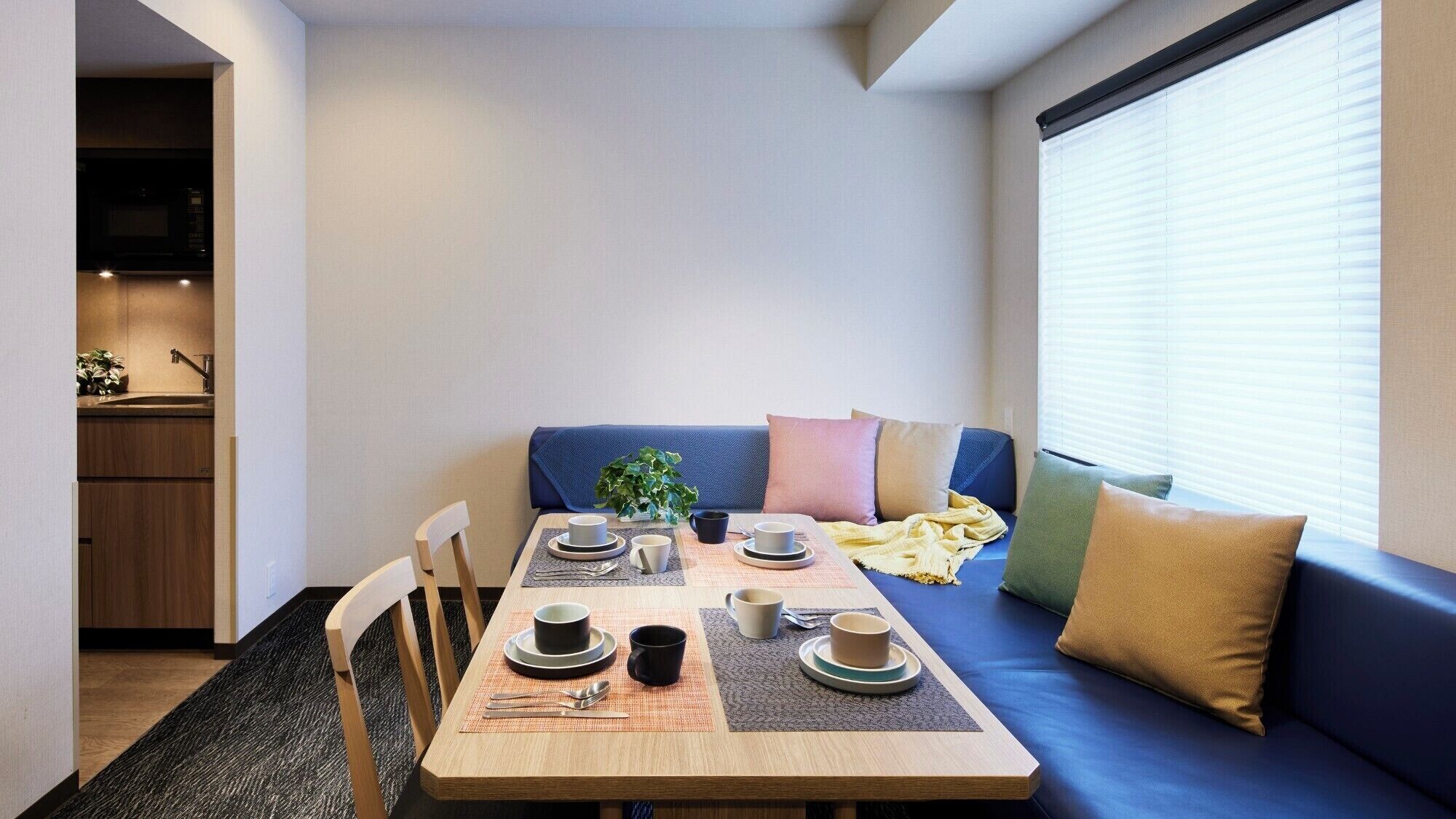 A compact living space with a dining table set for two, a sofa with cushions, a kitchen area with a microwave, and a window with blinds.