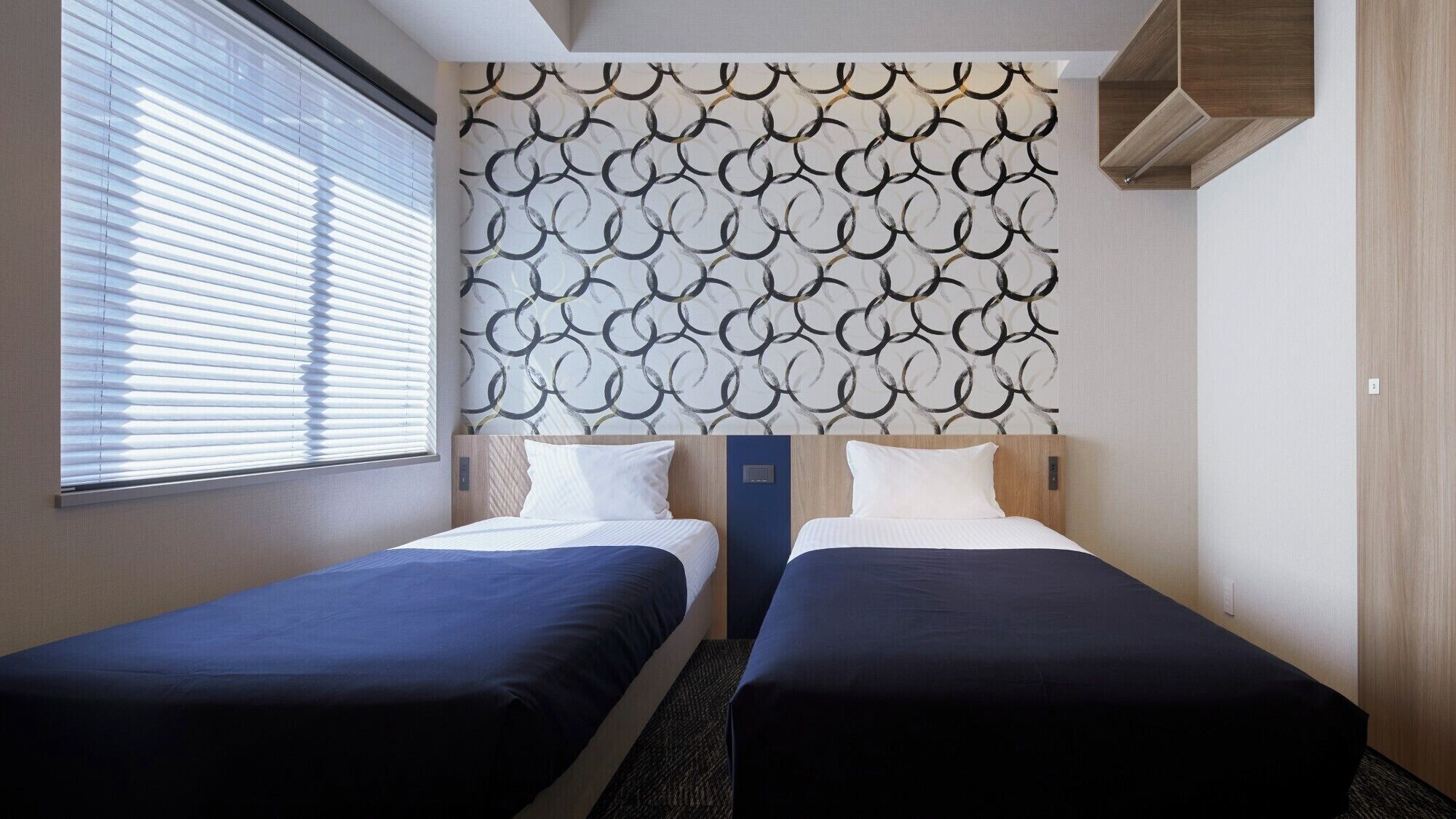 A hotel room with two beds, a patterned headboard, and a window with blinds.