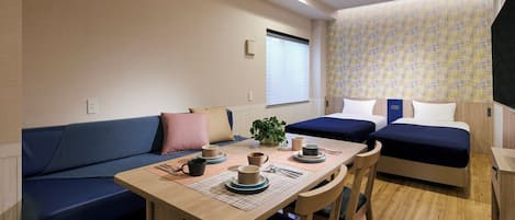 A compact hotel room with a sofa, two single beds, a small dining table with cups and a plant, and a patterned wall.