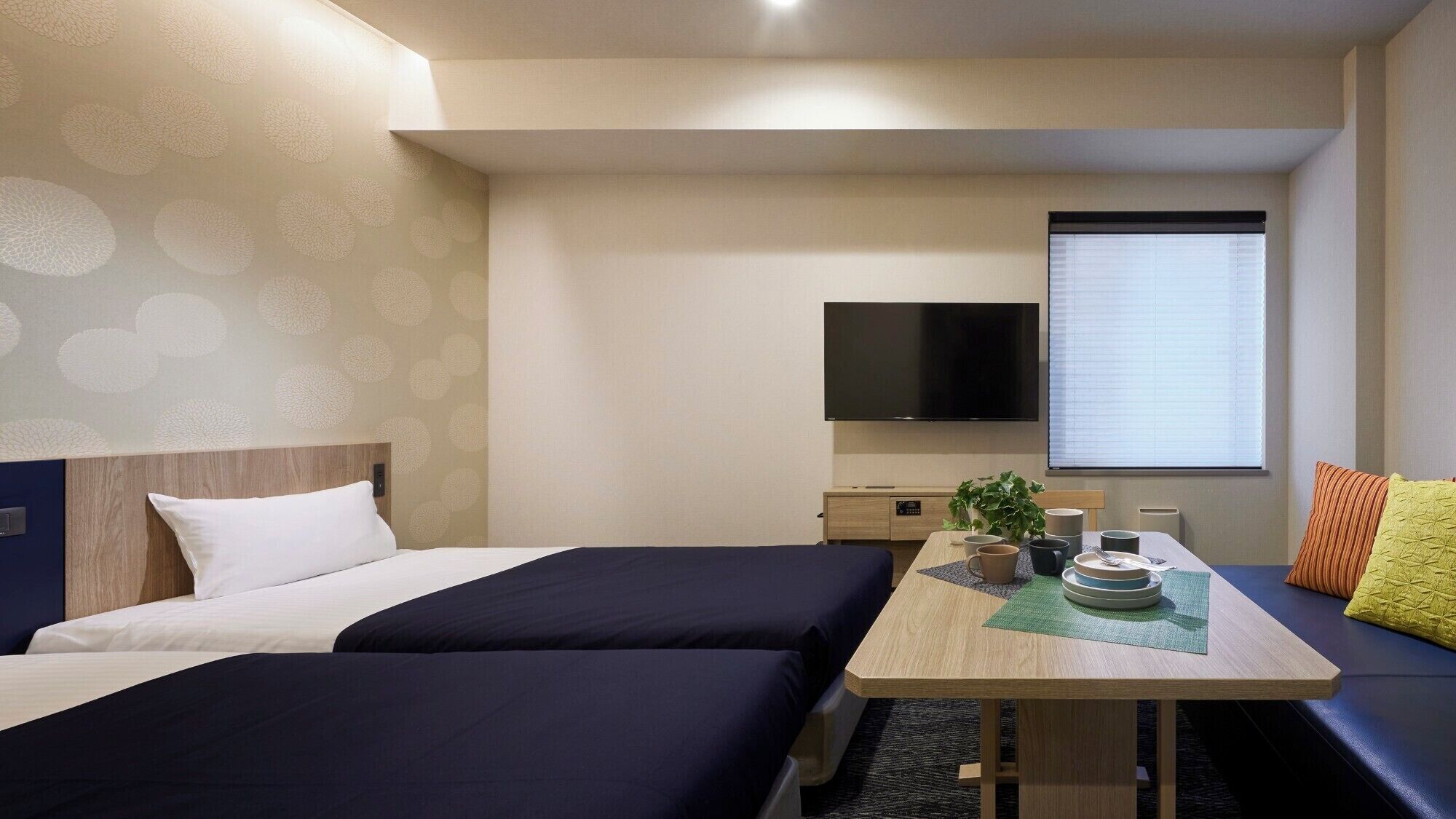Quarto de hotel com duas camas, uma televisão, uma mesa com uma planta e uma janela com persianas.