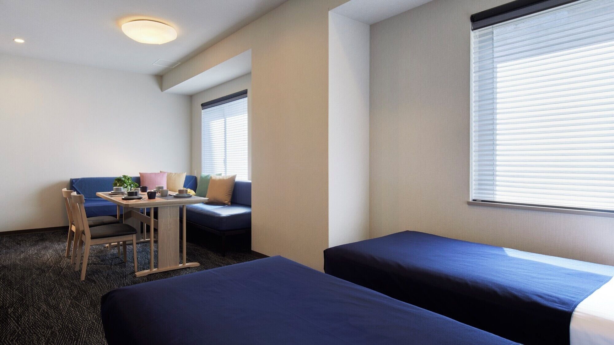 A hotel room with two single beds, a small table with a chair, a sofa, and a window with blinds.