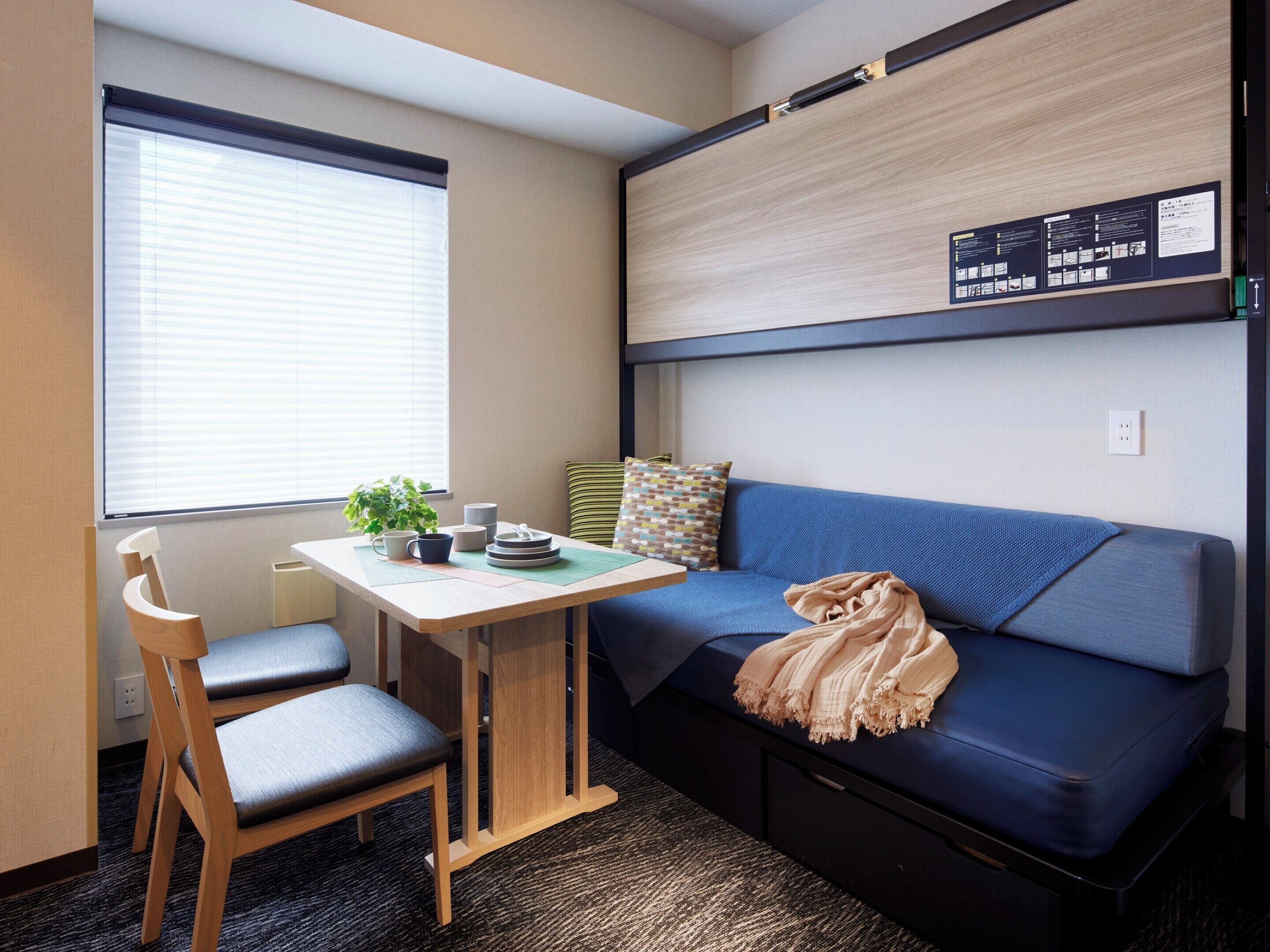Una habitación compacta con un sofá azul, una pequeña mesa de madera con una planta y una ventana con persianas.