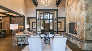 A spacious living room with a stone fireplace, a large chandelier, and a view of the outdoors through a large window. - Bound to Impress. Private Hot Tub, Golf Access & Exclusive Club Amenities! (Truckee)