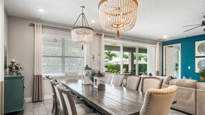 Un espace repas avec une longue table en bois, des chaises recouvertes d’un tissu beige et deux lustres.