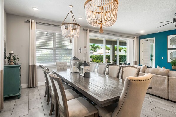 A dining area with a long wooden table, beige upholstered chairs, and two chandeliers. - Cinematic 7BR Encore with Home Theater & Pool (Kissimmee)