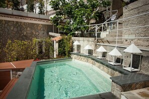 Ein Pool mit Steinwänden und einer Holzterrasse, umgeben von einer Steintreppe und Grünpflanzen.
