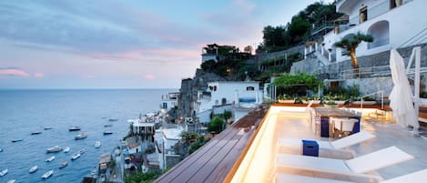 A coastal terrace with outdoor seating and a view of the sea and buildings.