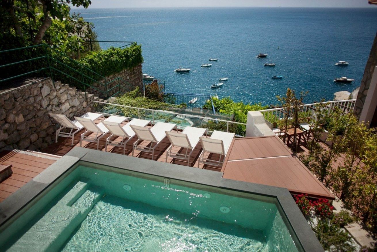 A pool area with lounge chairs overlooking the sea, a stone wall, and a wooden deck.