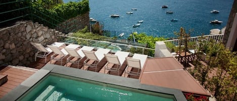 A pool area with lounge chairs overlooking the sea, a stone wall, and a wooden deck.