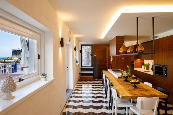 Une cuisine moderne avec un plan de travail en bois, des chaises blanches et une vue sur la mer grâce à une grande fenêtre.