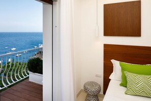 Une chambre à coucher avec une tête de lit en bois, un lit agrémenté d’oreillers verts et blancs, un rideau blanc et une vue sur la mer avec des bateaux.