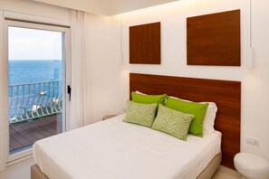 Une chambre d’hôtel avec un balcon donnant sur la mer, un lit agrémenté d’oreillers à motifs verts et blancs, et une tête de lit en bois.