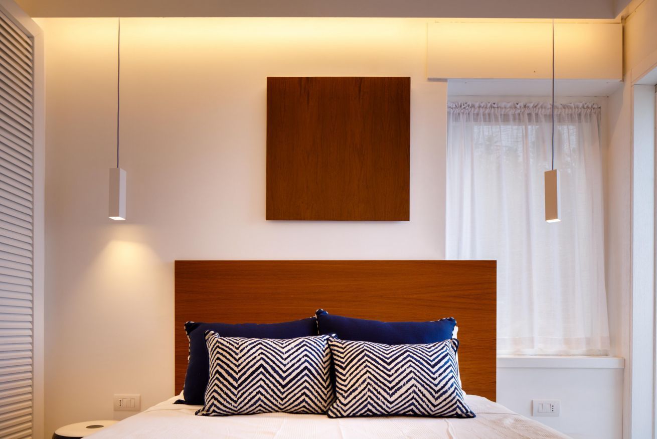 A modern bedroom with a wooden headboard, two patterned pillows, a bedside table, and a window with sheer curtains.