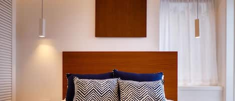 Un dormitorio moderno con cabecera de madera, dos almohadas estampadas, una mesita de noche y una ventana con cortinas traslúcidas.