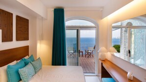 A bedroom with a bed, teal pillows, a wooden headboard, a window with a view of the sea, and a balcony with chairs. - Amalfi Coast Luxury Villa with Swimming Pool (Praiano)
