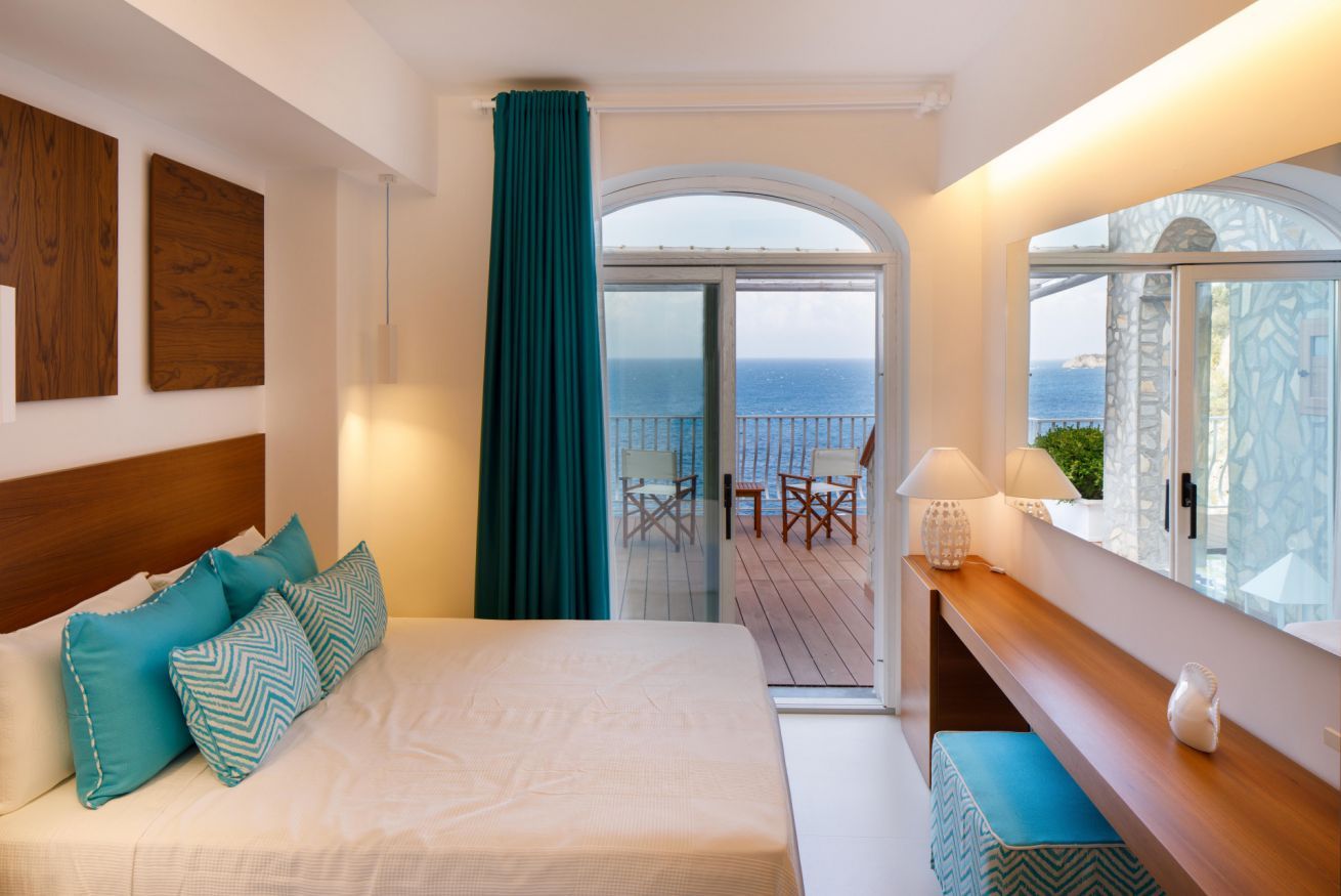 Une chambre à coucher avec un lit, des coussins de couleur turquoise, une tête de lit en bois, une fenêtre donnant sur la mer et un balcon avec des chaises.