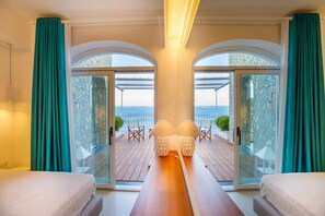 A hotel room with two beds, a wooden table with lamps, and a balcony with a view of the sea.